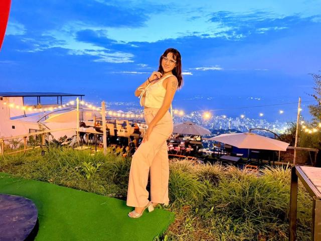 HanaJoud stands on green grass in a yellow dress, touching her neck, with blue evening sky and city lights in background. She has medium tits, long dark hair, and smiles.