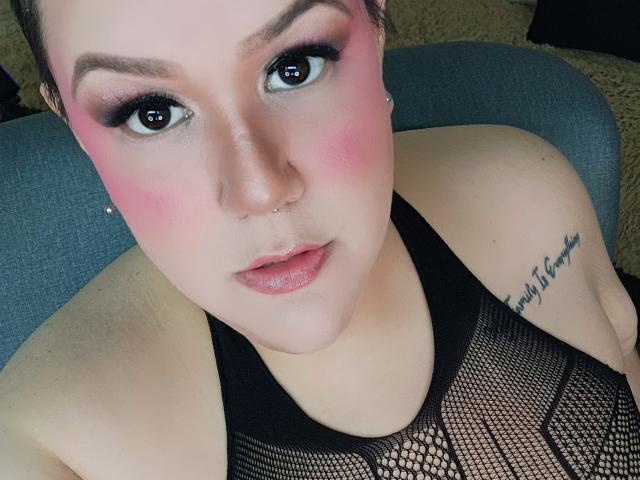 GiiannaMorrys with pink blush, black mesh top, nose piercing, tattoo on right shoulder, and short brown hair, looking at camera with slightly parted pink lips.