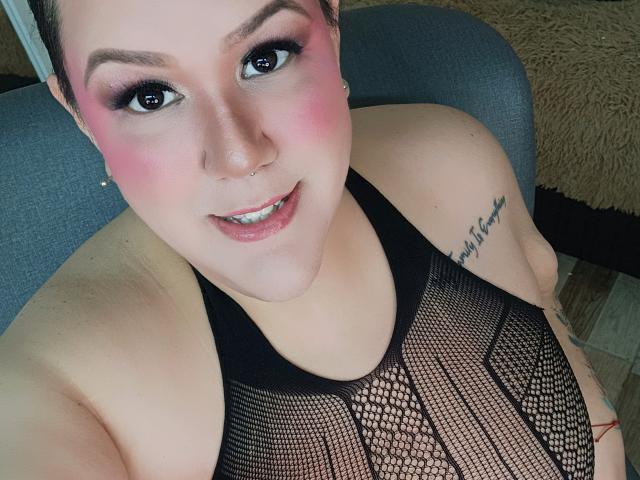 GiiannaMorrys with pink eyeshadow, nose ring, and black mesh top, showing her tits and tattoo on right shoulder, smiling at the camera.