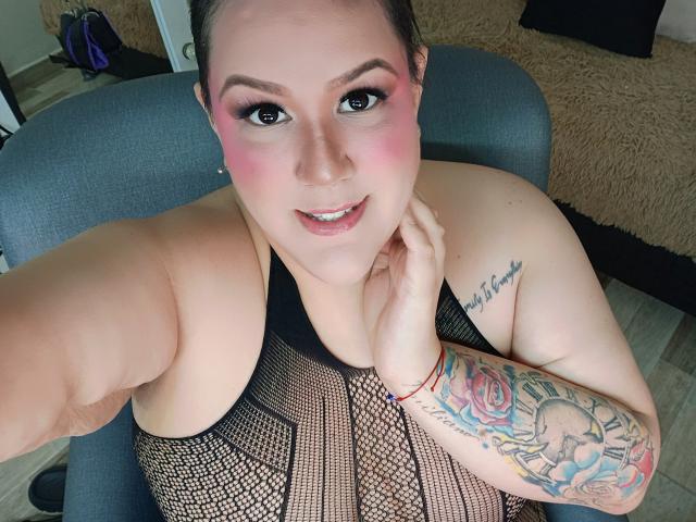 GiiannaMorrys with pink eye makeup, smiling, showing tattoos on her arm, wearing a black mesh top, holding her chin, and taking a selfie. Her tits are visible.
