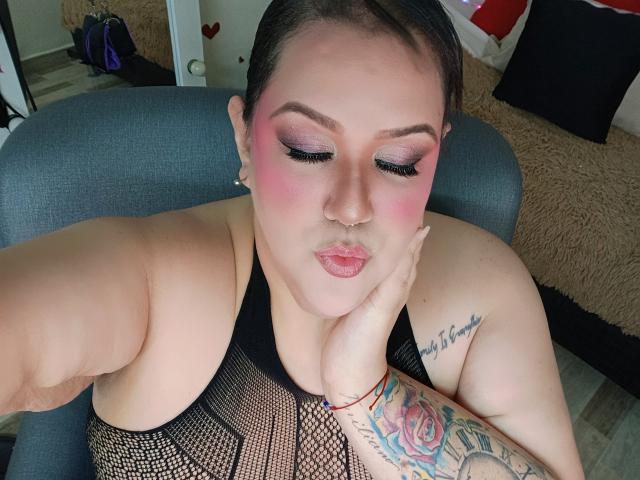 GiiannaMorrys with pink eyeshadow, pouty lips, black mesh top, tattooed arm, nose ring, and red bracelet, sitting in chair, eyes closed, hand on cheek.