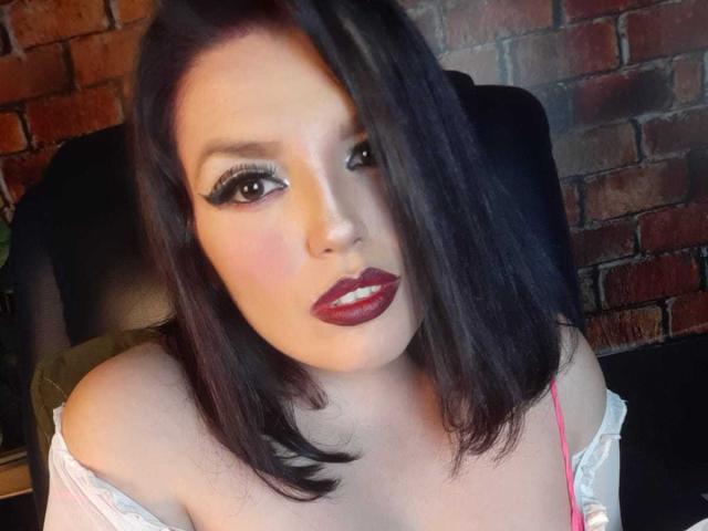 SachyMoon has dark, shoulder-length hair, heavy eyeliner, and red lipstick. She's looking at the camera with a slightly open mouth, wearing a pink strap, and off-shoulder white top.