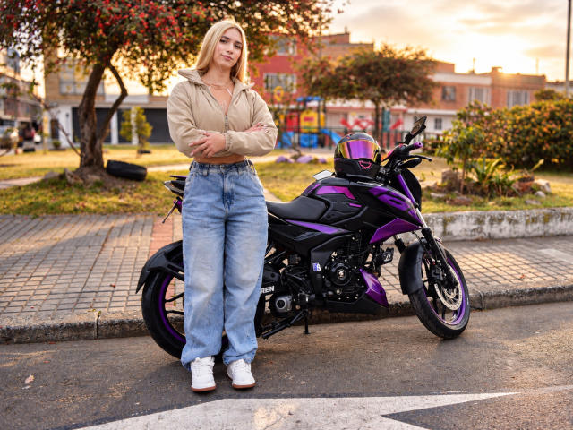 TanniaMoon standing with arms crossed, wearing a beige shirt, blue jeans, and white shoes, next to a purple motorcycle with a matching helmet, in a sunny urban setting.