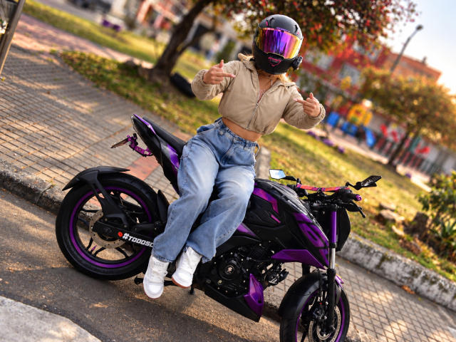 TanniaMoon sentada en una motocicleta morada, vistiendo una sudadera beige, jeans azules de cintura alta, zapatillas blancas y un casco reflectante, levantando el pulgar con ambas manos..