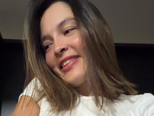 Gabrielafoxxy's face is smiling with slightly parted red lips, brown eyes, and shoulder-length brown hair, wearing a white shirt, with a small mole on her right cheek.