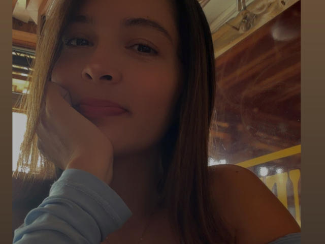 Gabrielafoxxy hat langes, braunes Haar, ein süßes Gesicht und legt ihr Kinn in ihre Hand, wobei sie leicht lächelt. Sie trägt ein blaues Hemd, und es ist ein Selfie..
