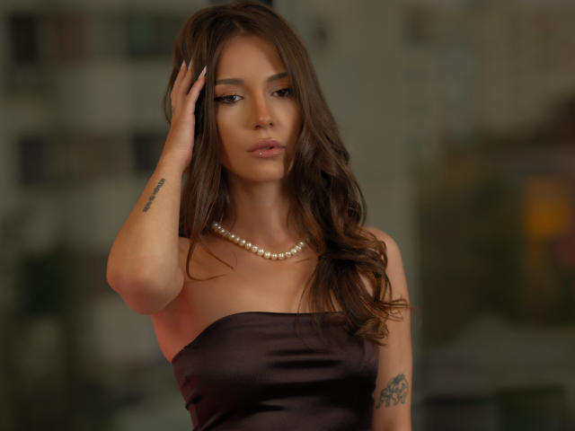 LexiraOnen with wavy brown hair, wearing a strapless black dress and pearl necklace, hand on head, tattoo on left arm, looking serious, tits visible, soft light.
