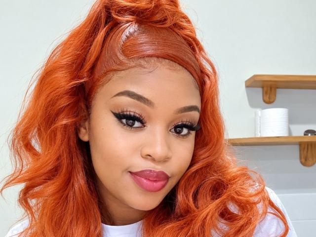 ThiqueKitty, avec ses cheveux orange vif, ses longs cils et son rouge à lèvres rose, sourit à l'appareil photo. Ses fesses ne sont pas visibles, seulement son visage et le haut de son corps..