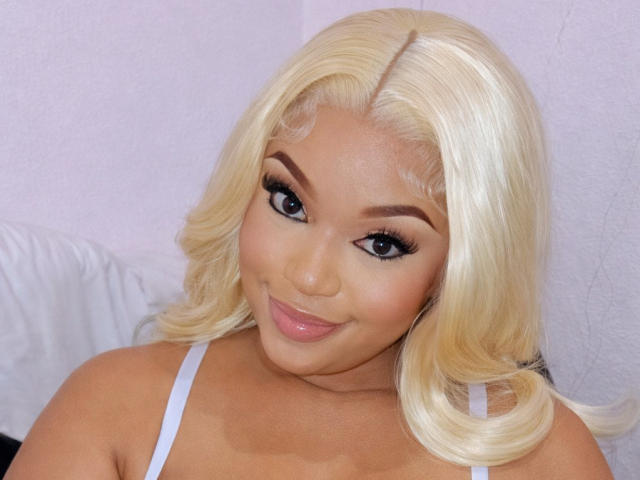 ThiqueKitty a les cheveux blonds, des sourcils épais, de longs cils noirs, et elle sourit avec des lèvres roses et brillantes. Elle porte un haut blanc, et ses seins sont petits..