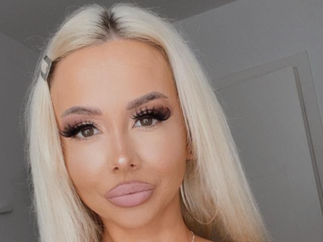 MoniqueBliss has blonde hair, green eyes, and full pink lips with heavy black eyeliner and long lashes. She's wearing a hair clip on the left side of her head.