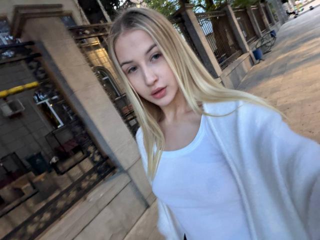 Blonde woman with long hair, wearing a white shirt and open jacket, standing on a sidewalk, taking a selfie, showing her tits, with a neutral expression.