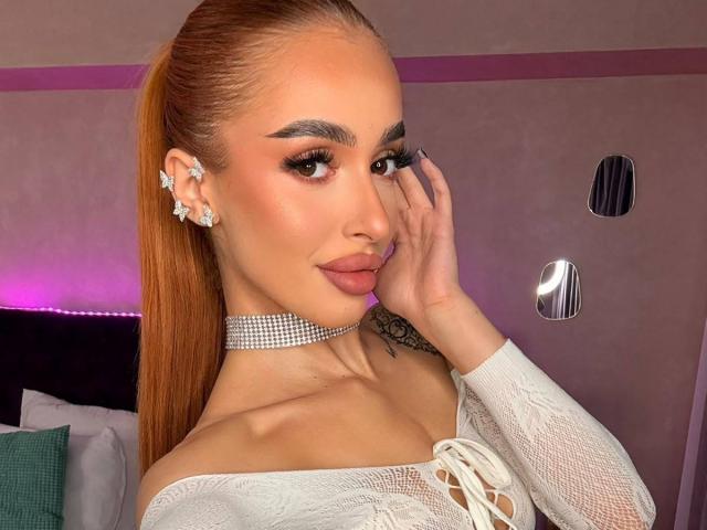 MarissaFex has orange hair in a ponytail, wearing a white lace off-shoulder top, diamond earrings, and a diamond choker, with her hand on her face, showing off her tits.