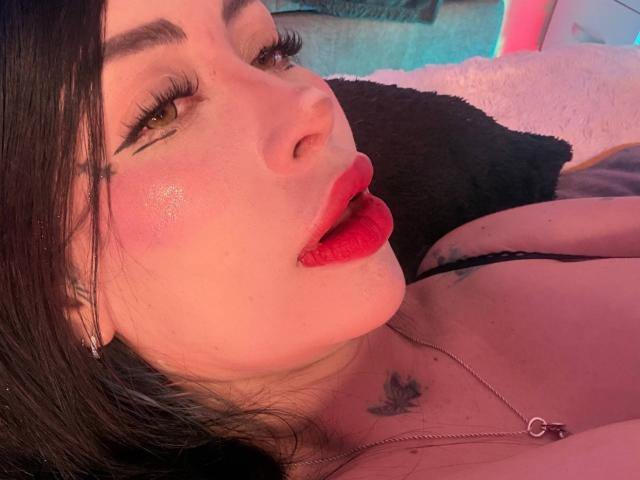 Kkole's face with red lipstick, black eyeliner, and shiny pink cheeks. She has tattoos, a black strap, and a necklace, lying on a black pillow. Kkole's tits are visible.