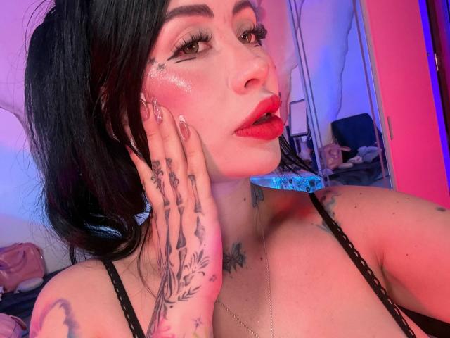 Kkole with black hair, red lipstick, and tattoos on hands and chest, touching face, wearing black lace top, glowing skin, blue light, and pink light in background.