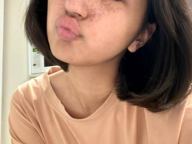HeikeCarchi with puffy pink lips, freckled nose, and short brown hair, wearing a peach shirt, pouting in a selfie. Her small tits are slightly visible.