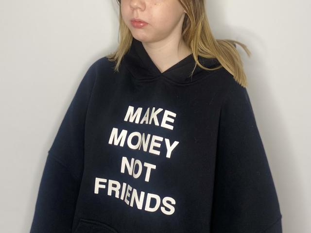 LauranTanton, com pele clara e cabelo loiro, vestindo um moletom preto com a frase "MAKE MOVIES NOT FRIENDS", tem uma expressão neutra e está em pé em frente a uma parede lisa..