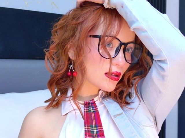 Emily Klein heeft rood krullend haar, een zwarte bril, rode oorbellen, een witte blouse, een rode geruite stropdas en een grijze jas, met haar arm omhoog en haar hand in haar haar..