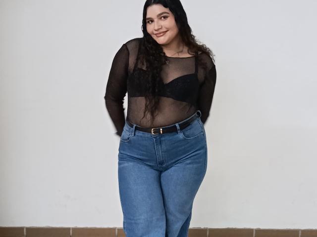 ChubbyKinkyWoman with long curly black hair, wearing a sheer black top showing small tits, blue high-waisted jeans, and a black belt, standing and smiling.