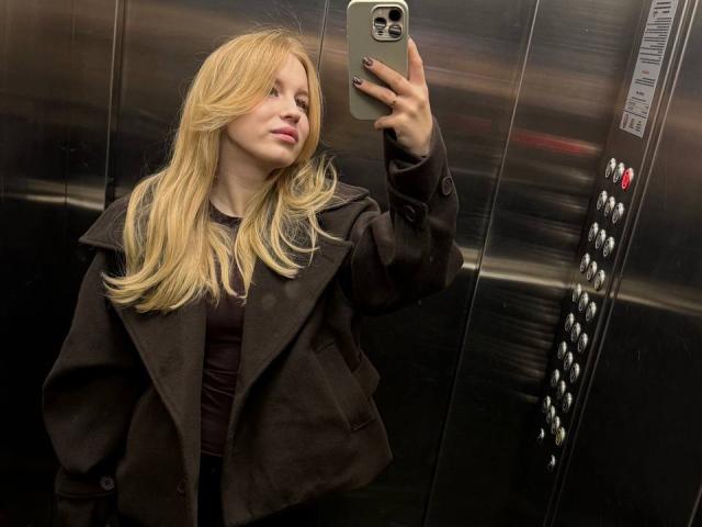 MariluChouinard with long blonde hair, brown coat, and black shirt, taking a selfie in a metal elevator, holding a green phone, with black nails.
