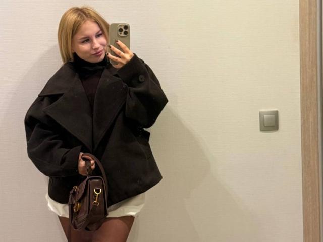 MariluChouinard with blonde hair, black coat, white shorts, holding brown bag, phone in hand, smiling, standing, visible butt, taking mirror selfie.
