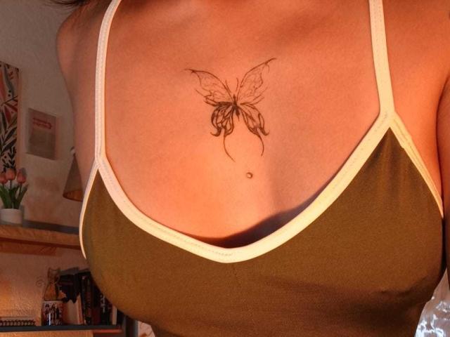 MilaThorne's tits, showing a delicate butterfly tattoo on her chest, wearing a green tank top with white trim, sunlit skin, and a small dot below the tattoo.