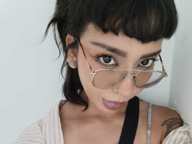 MilaThorne with dark hair, glasses, and pink lipstick, pouting slightly, wearing a striped shirt, black strap, and visible tattoo on right shoulder.