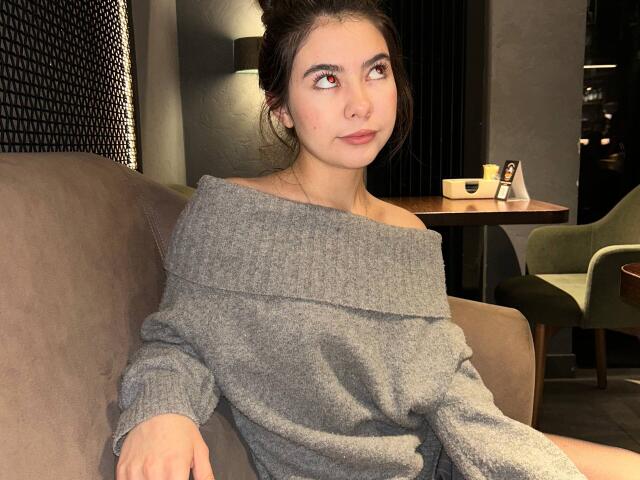 AnneKofron with dark hair in a bun, wearing an off-shoulder gray sweater, sitting on a brown couch, looking to the side with a slight smile, in a dimly lit room.