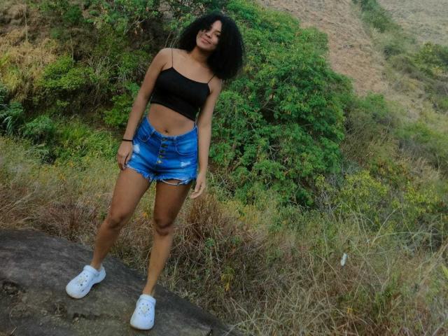 HornySweetEbony with curly black hair, black crop top, blue denim shorts, and white sneakers, standing on a rock, looking down, outdoors, green bush, brown grass, sunny day.