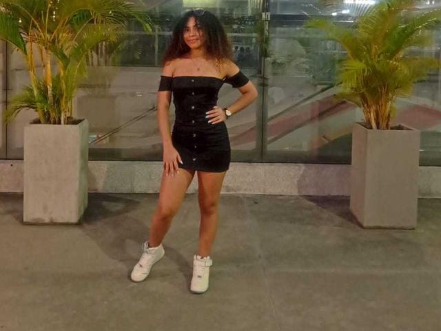 HornySweetEbony with curly brown hair, wearing a black off-shoulder dress, buttoned down, and white boots, standing in front of two potted plants, smiling.