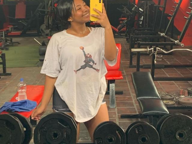 DreamingOfU draagt een wit shirt met een bedrukte afbeelding, ze glimlacht en maakt een spiegelzelfportret in de sportschool, ze draagt geen broek, toont haar achterwerk, en heeft een gele telefoon bij zich..