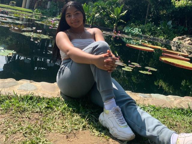 DreamingOfU with tan skin, black hair, small tits, wearing a strapless top, gray pants, and white sneakers, sitting by a lily pond, legs crossed, smiling.