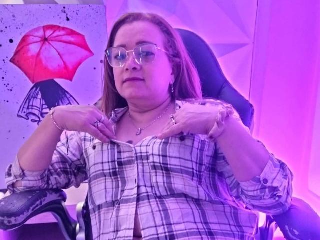 SammiraMorgan has glasses, red hair, and is wearing a plaid shirt with buttons undone, exposing her tits. She's sitting in a chair with hands on her chest.