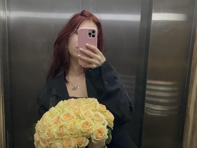 AlexXas met rood haar, een zwart shirt, die een boeket gele en oranje rozen vasthoudt, maakt een spiegel-selfie in een metalen lift, met een telefoon in een roze hoes die zijn gezicht bedekt..