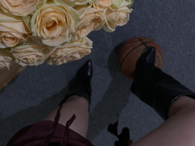 AlexXas's legs in black boots with pointy toes, wearing a red skirt, standing next to a basketball on a gray carpet, and a vase of beige roses.