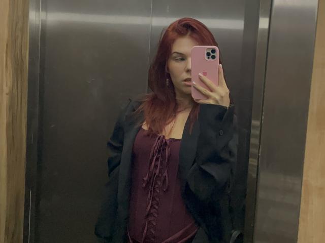 AlexXas with red hair, wearing a maroon top and black jacket, taking a mirror selfie in an elevator, holding a pink phone, showing her tits slightly visible.