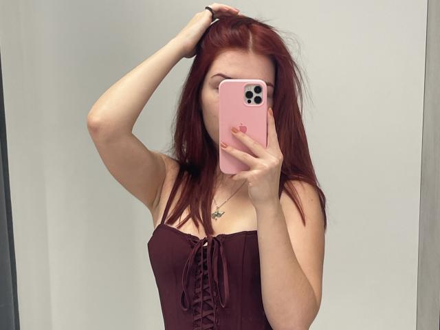 AlexXas with red hair, wearing a strapless maroon top, holding a pink phone, covering her face, raising her right hand through her hair, showing her tits.
