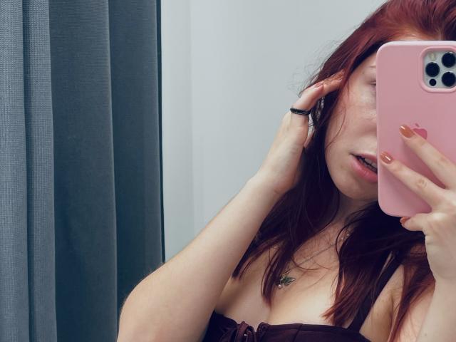 AlexXas met rood haar, die een roze telefoon vasthoudt, laat haar decolleté zien in een top zonder bandjes, heeft een vinger in haar haar, draagt een kleine ketting en heeft gouden nagellak..