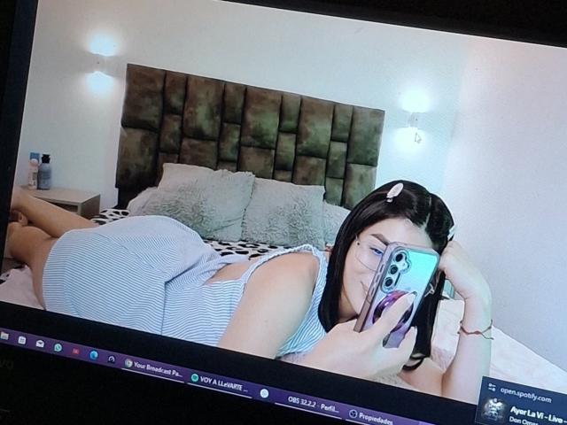PaulhettJoens lying on bed, striped dress, butt up, taking mirror selfie with phone, black hair, glasses, white wall, green headboard, pillows, water bottle on nightstand.