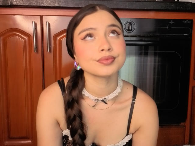 VictoriaKyros with pouty lips, looking up, has a braided ponytail, black lace top, white choker, and colorful earrings, standing in front of a wooden kitchen cabinet.
