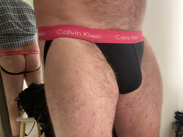 OfficeBitcyh's hairy, hairy-legged, hairy-pelvis, black Calvin Klein briefs with red waistband, showing his black-ass in mirror reflection. Plaid shirt, white wall, black chair in background.