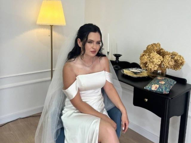 Charlette Biskach est assise sur une chaise, vêtue d'une robe blanche à une épaule avec un voile, ses seins étant légèrement visibles, dans une pièce avec une table noire et des fleurs dorées..