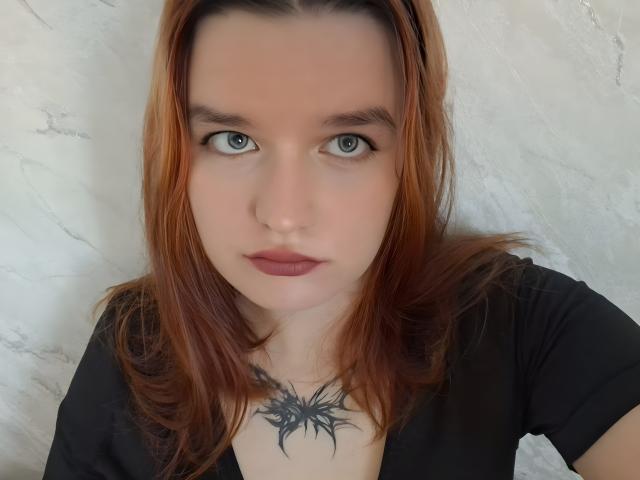 Jacquiline Drumbore tem cabelo ruivo, olhos azuis e uma tatuagem preta no peito. Ela está vestindo uma camisa preta, e seus seios estão parcialmente visíveis..