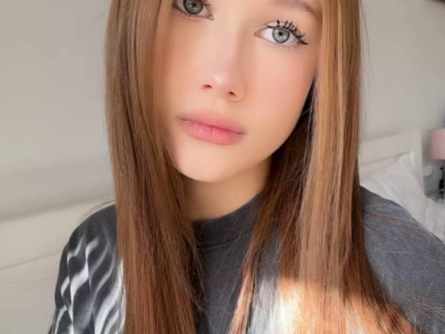 RainaSpotorno has long, straight brown hair, light green eyes, and pink lips. She's wearing a gray shirt, and her eyelashes are long and dark.