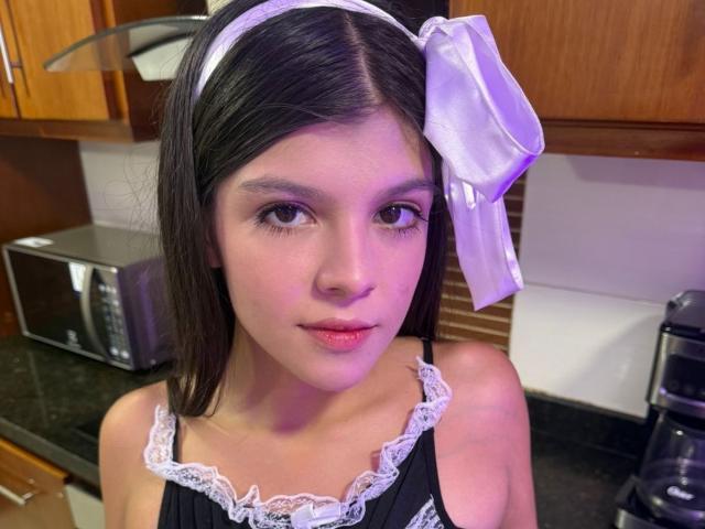 RilleyFoyx has straight black hair, a white lace headband with a purple ribbon, and a black lace-trimmed top, showing her small, cute tits in a kitchen.