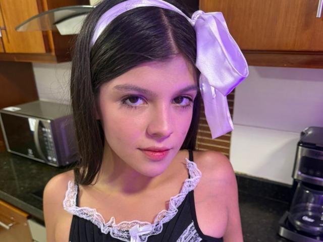 RilleyFoyx with dark hair, white headband, and large white bow, wearing a black lace-trimmed top, has small tits and a cute face, in a kitchen.