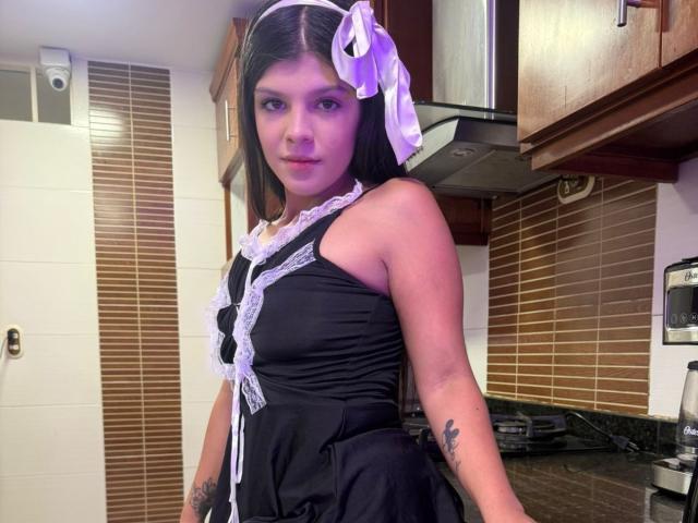RilleyFoyx, wearing a black maid outfit with white lace, purple hair bow, tattoos on arms, standing in kitchen, purple makeup, black dress, looking at camera, medium shot.