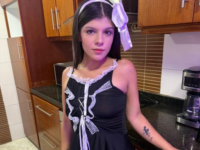 RilleyFoyx with long black hair, white headband, black lace-up dress, white ribbons, small tits, tattoo on right arm, standing in kitchen, looking at camera, slight purple lighting.