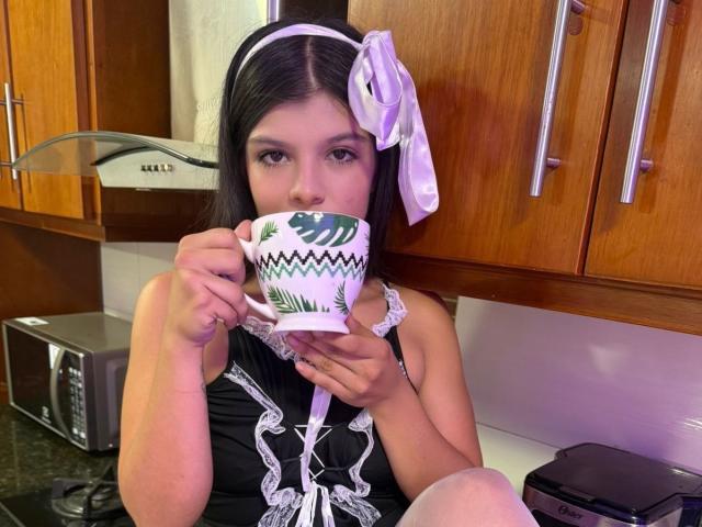 RilleyFoyx with black hair, white headband, purple ribbon, black lace-up dress, holding green-patterned cup, sipping, in wooden kitchen, wearing white lace stockings, visible knees.