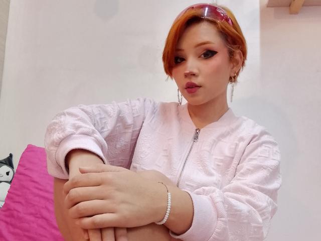 ChloeeWilliams with orange hair, pink eyeshadow, and pink lipstick, wearing a pink, textured, zip-up shirt, arms crossed, looking at camera, bracelet on wrist.