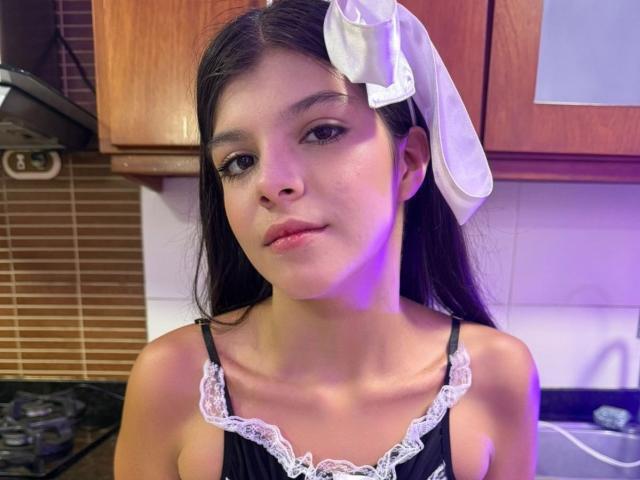 RilleyFoyx with long black hair, white bow, wearing a black lace-trimmed top, standing in a kitchen, looking at the camera with a slight smile. Purple light on right.